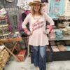 Woman wearing a cowboy hat and fringe jeans in an clothing store setting wearing a graphic t that says "all american sweetheart"