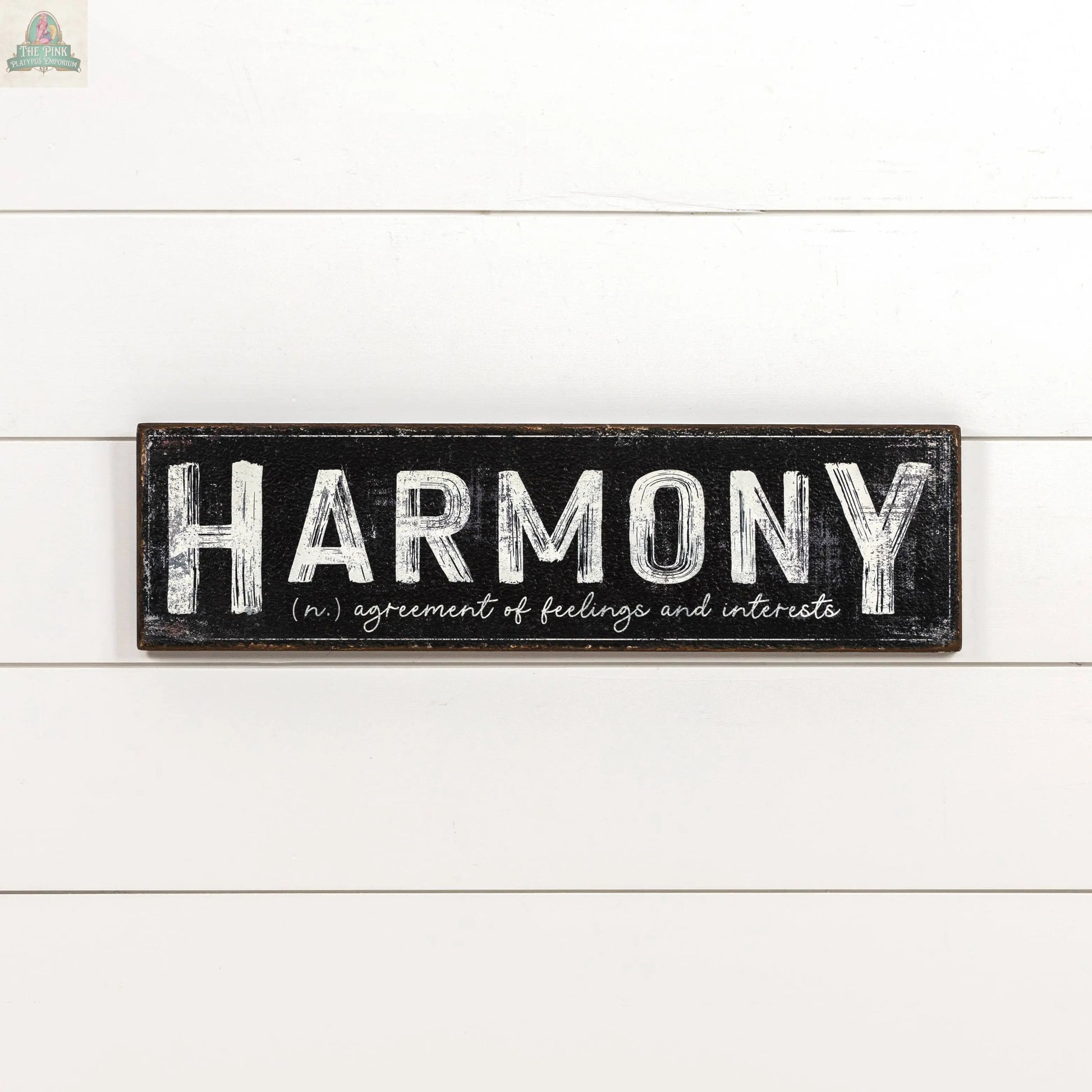 The 24" HARMONY METAL SIGN by Ragon House is a black rectangular metal sign with bold white "HARMONY" lettering and its meaning below, adding an elegant accent to your home decor.
