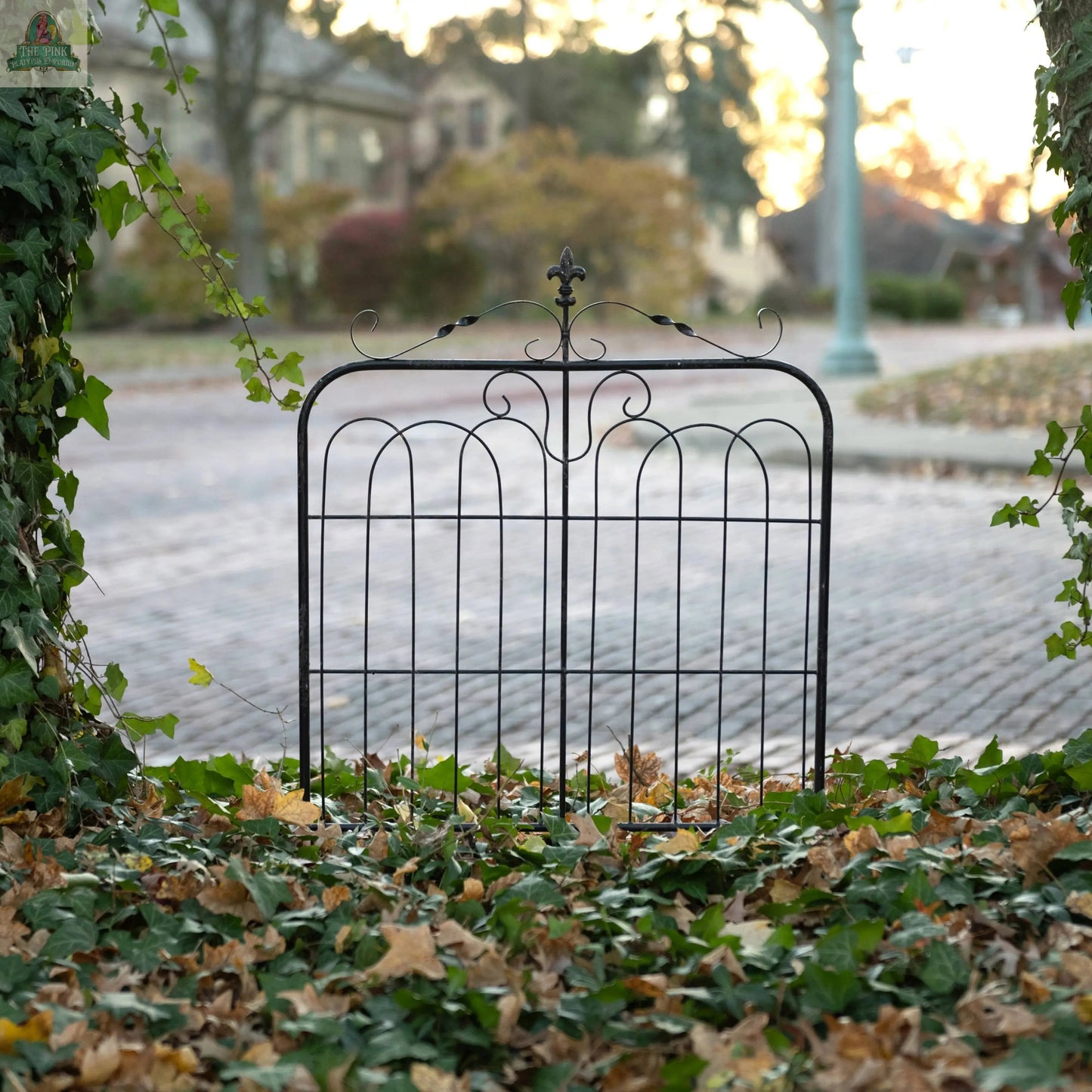 The 32" AGED BLACK GARDEN GATE stands among fallen leaves and ivy, with a brick-paved street and soft-focus houses in the background at sunset.