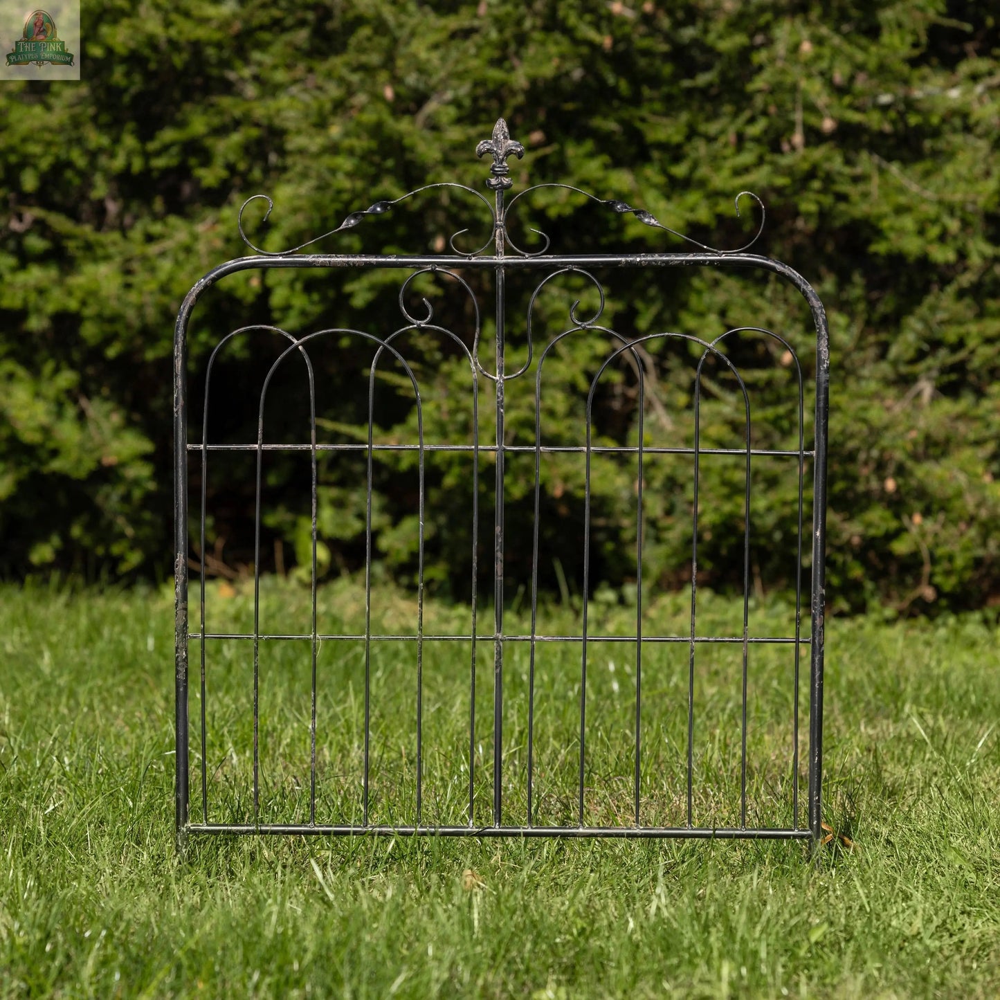 The 32" AGED BLACK GARDEN GATE, featuring ornate scrollwork, stands on a grassy lawn with dense green trees in the background under bright sunlight.