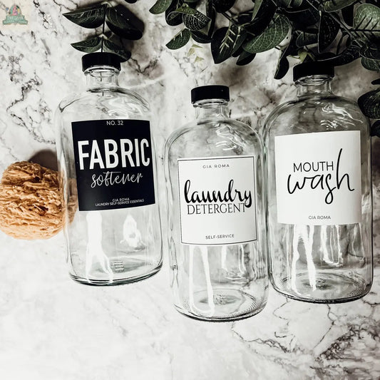 Three 32oz Glass Clear dispenser bottles labeled Fabric Softener, Laundry Detergent, and Mouth Wash sit on a marble surface with a natural sponge, green foliage, and waterproof labels for easy identification.