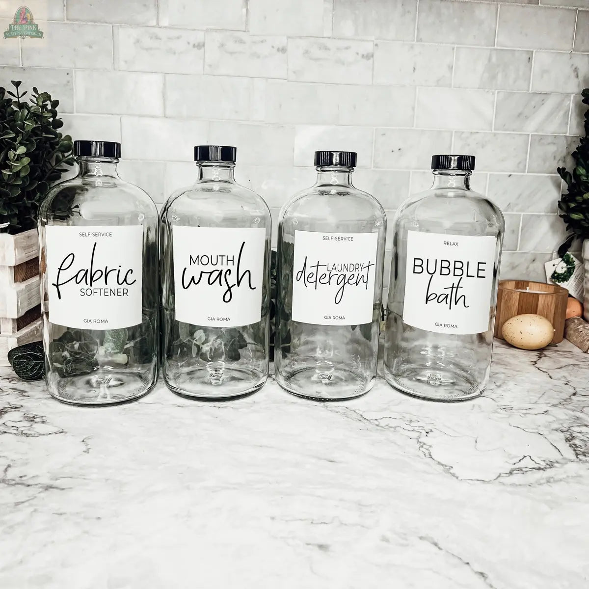 Four 32oz Glass Clear bottles with black caps, labeled for Fabric Softener, Mouth Wash, Laundry Detergent, and Bubble Bath, sit on a marble countertop with plants and kitchen items in the background. Waterproof labels included.