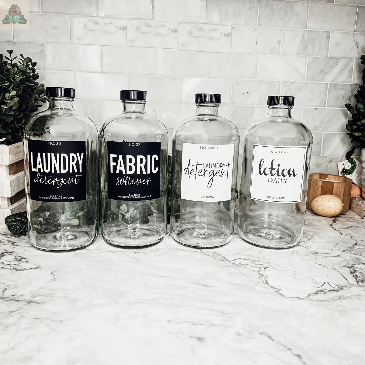 Four 32oz Glass Clear dispenser bottles with black pumps and waterproof labels for Laundry Detergent, Fabric Softener, and Lotion Daily are arranged on a marble countertop, surrounded by greenery and kitchen accents.