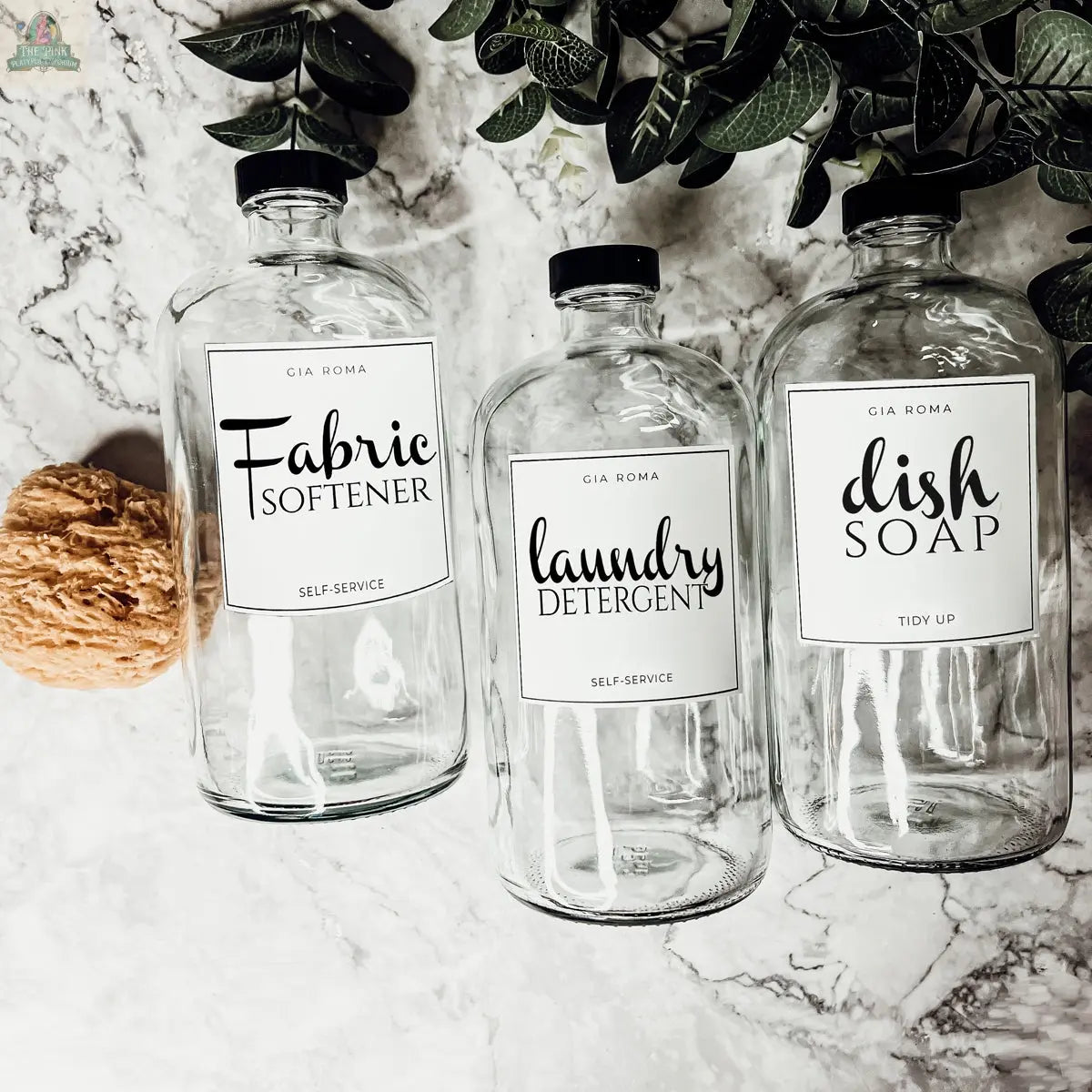 Three 32oz Glass Clear dispenser bottles labeled Fabric Softener, Laundry Detergent, and Dish Soap are arranged on a marble surface with a natural sponge and green leaves, each featuring waterproof labels for reusable organization.