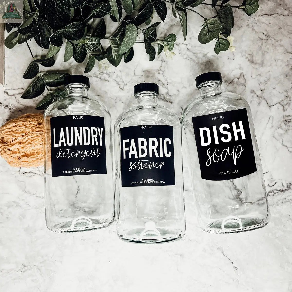Three empty 32oz Glass Clear dispenser bottles labeled Laundry Detergent, Fabric Softener, and Dish Soap sit on marble with a natural sponge and green leaves in the background, each featuring a waterproof label.