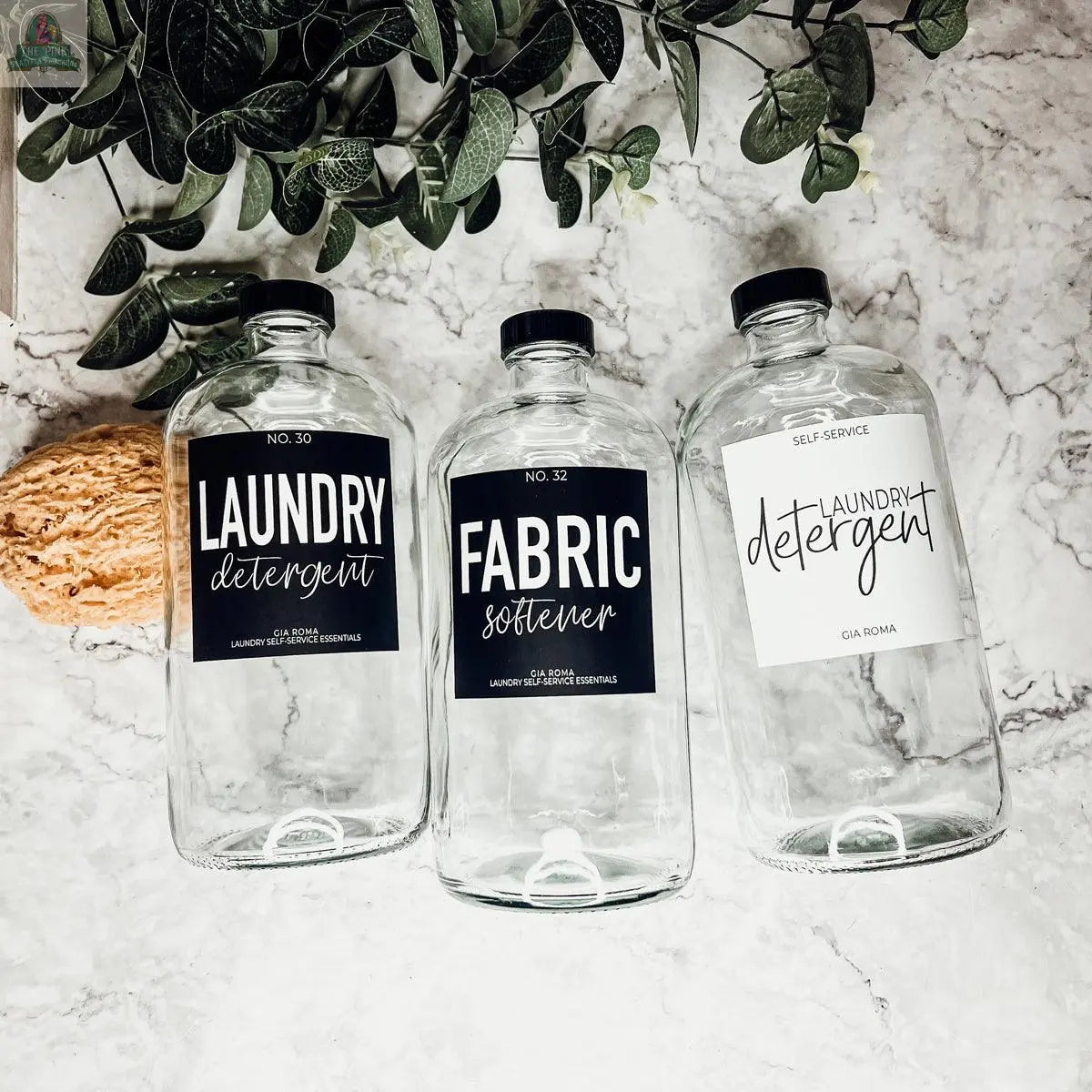 Three 32oz Glass Clear bottles labeled "Laundry Detergent," "Fabric Softener," and "Laundry Detergent" sit on a marble surface near a plant and sponge, making them ideal reusable dispensers for your laundry room.