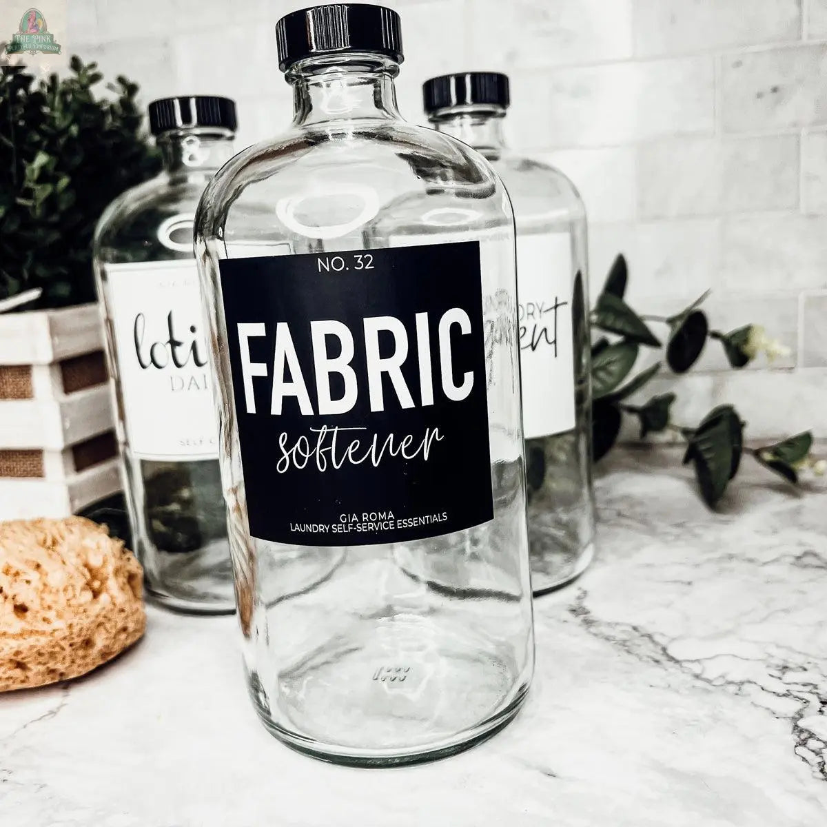 A 32oz Glass Clear dispenser labeled FABRIC softener sits on a marble counter. In the background, reusable glass bottles labeled lotion and laundry are visible, next to a plant and natural sponge. All bottles have waterproof labels.