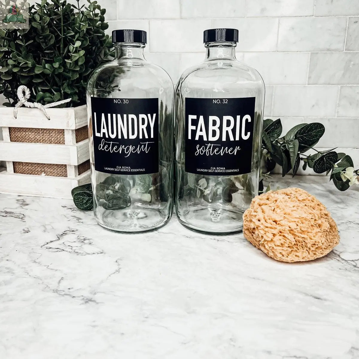 Two 32oz Glass Clear dispenser bottles labeled Laundry Detergent and Fabric Softener sit on a marble countertop beside a yellow sponge, with a small decorative plant in the background.