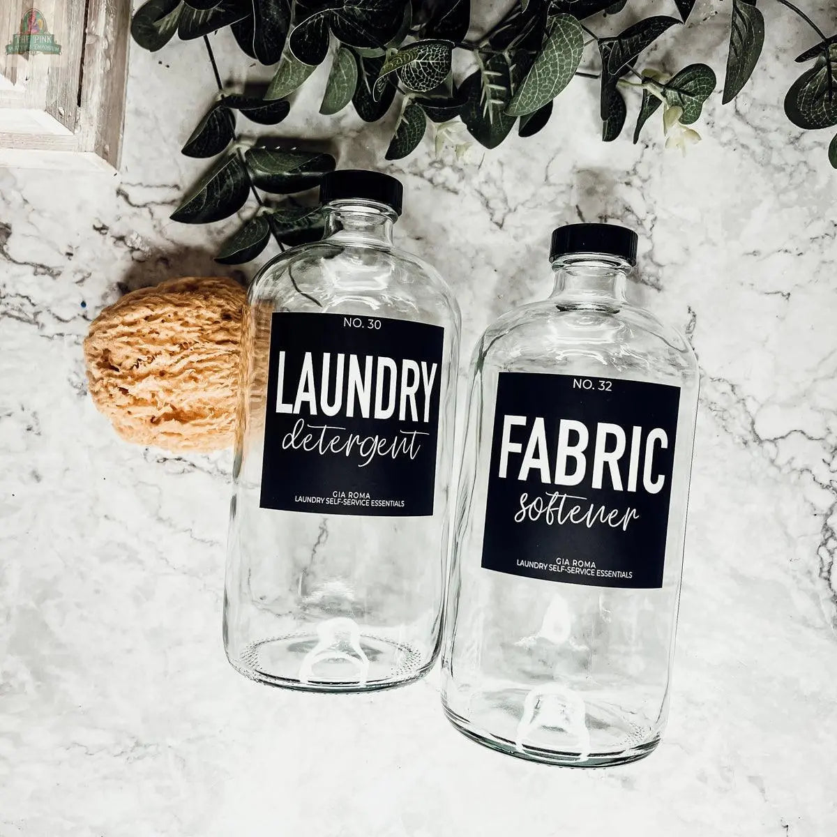 Two 32oz Glass Clear dispenser bottles labeled Laundry Detergent and Fabric Softener sit on white marble with a yellow sponge and green plant, featuring stylish waterproof labels for reusable glass bottles.