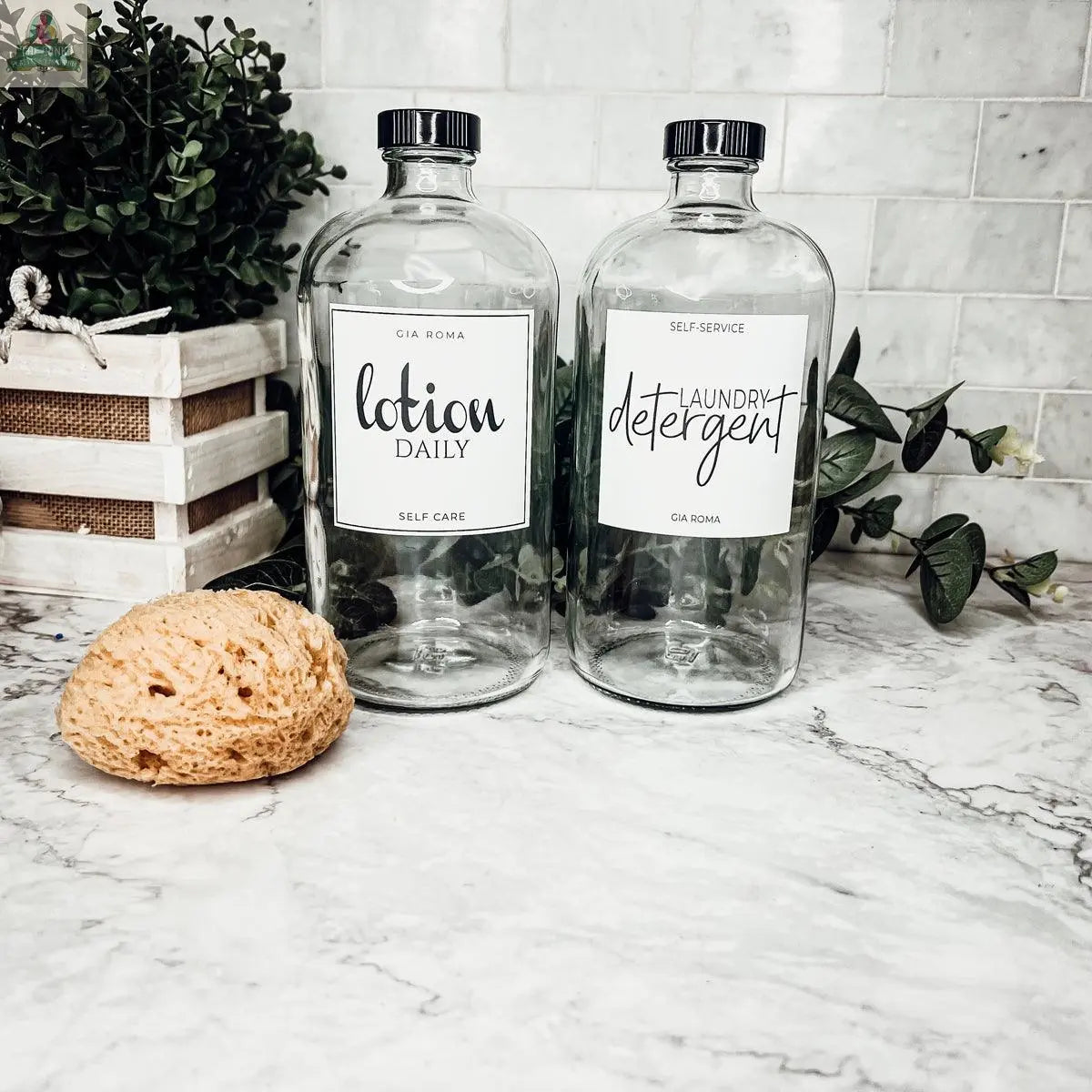 Two 32oz Glass Clear bottles with waterproof labels—lotion daily and laundry detergent—sit on a marble counter. A yellow sponge, small potted plant, and leafy vines add a fresh touch to the scene.