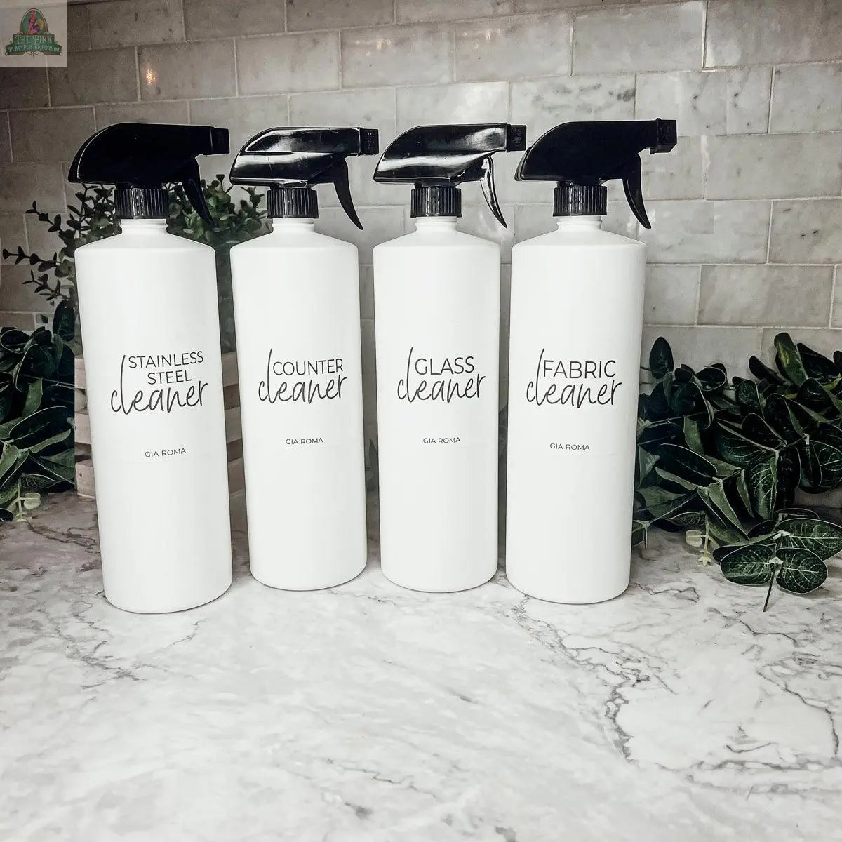 Four 32oz Plastic Cleaning Bottles labeled Stainless Steel Cleaner, Counter Cleaner, Glass Cleaner, and Fabric Cleaner sit on a marble countertop with green plants and white subway tile, each with a waterproof label.