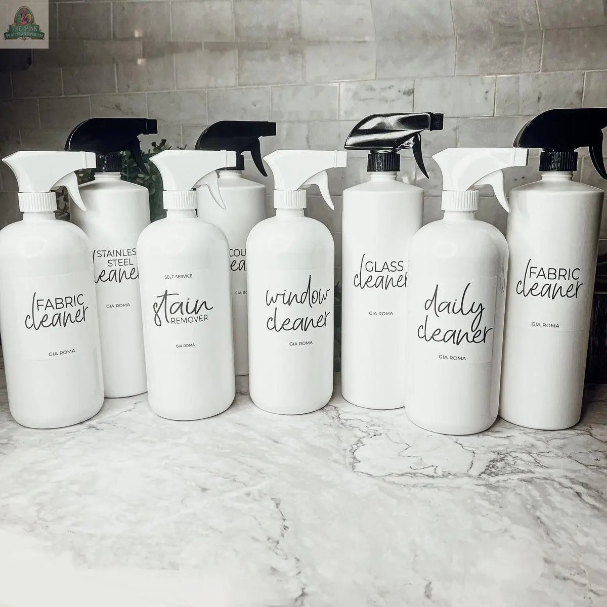 A row of eight 32oz Plastic Cleaning Bottles with black and white waterproof labels displays various cleaners—fabric, stainless steel, stain remover, window, and glass—neatly arranged on a kitchen counter.