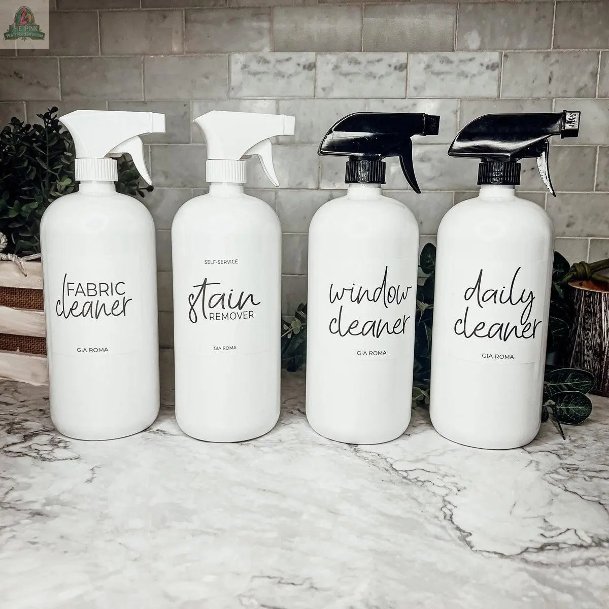 Four 32oz Plastic Cleaning Bottles with waterproof “Fabric Cleaner,” “Stain Remover,” “Window Cleaner,” and “Daily Cleaner” labels feature black and white nozzles. These refillable bottles sit on a marble counter near tile and green plants.