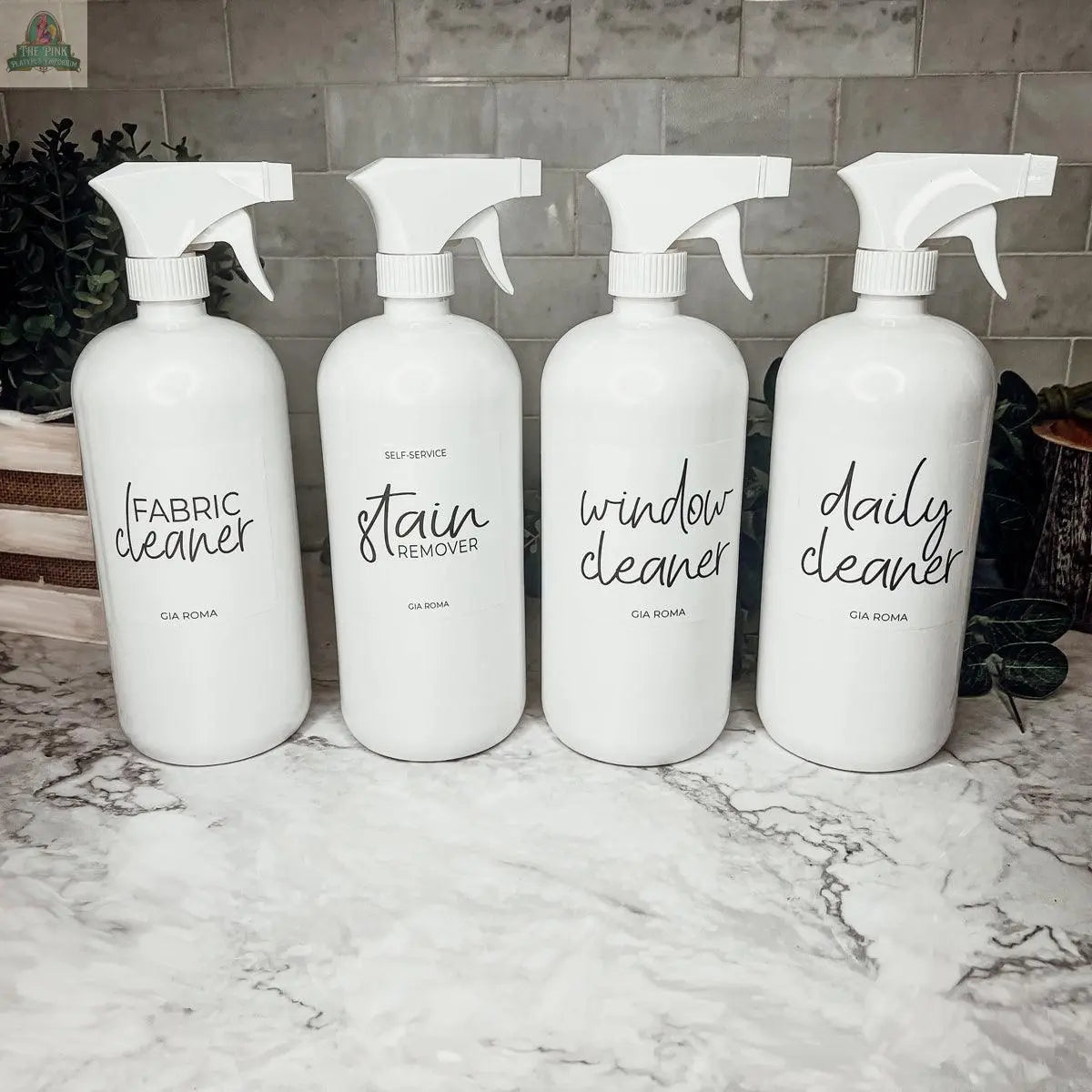 Four 32oz Plastic Cleaning Bottles labeled fabric cleaner, stain remover, window cleaner, and daily cleaner sit side by side on a marble countertop with a tiled backsplash and greenery. Each refillable bottle features a waterproof label.