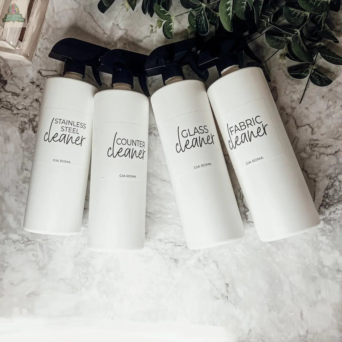 Four 32oz Plastic Cleaning Bottles—labeled stainless steel cleaner, counter cleaner, glass cleaner, and fabric cleaner—sit on a marble surface with waterproof labels. Green leaves rest in the top right corner.