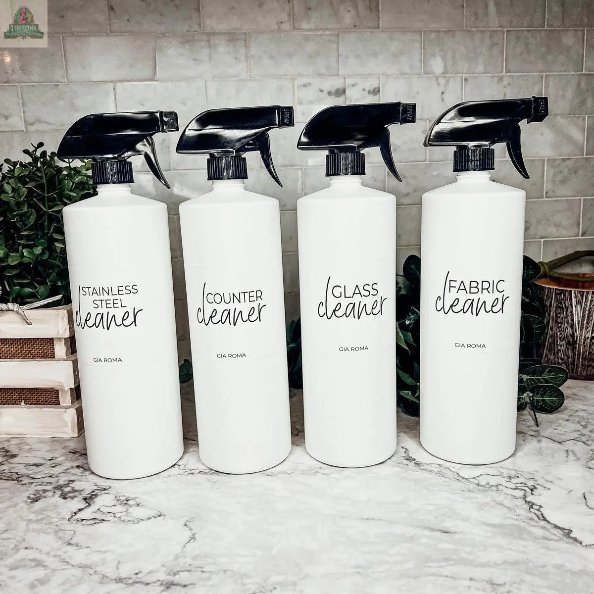 Four 32oz Plastic Cleaning Bottles labeled Stainless Steel Cleaner, Counter Cleaner, Glass Cleaner, and Fabric Cleaner sit on a marble countertop with waterproof labels, in front of a tiled backsplash and green plants.