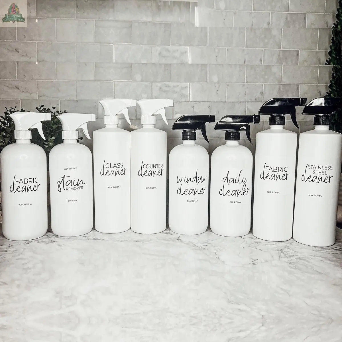 Eight 32oz Plastic Cleaning Bottles with black and clear nozzles are lined up on a marble countertop. Each refillable bottle has a waterproof label for fabric, glass, counter, window, daily, and stainless steel cleaning.