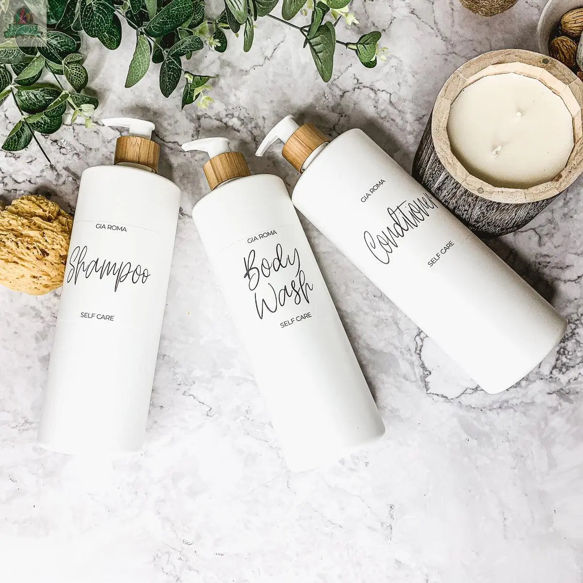 Three 32oz Plastic White Dispensers labeled shampoo, body wash, and conditioner sit on a marble surface with a candle, greenery, and a natural sponge nearby, creating a clean, spa-like setting.