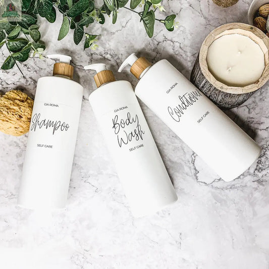 Three 32oz Plastic White Dispensers labeled shampoo, body wash, and conditioner sit on a marble surface with a candle, greenery, and a natural sponge nearby, creating a clean, spa-like setting.