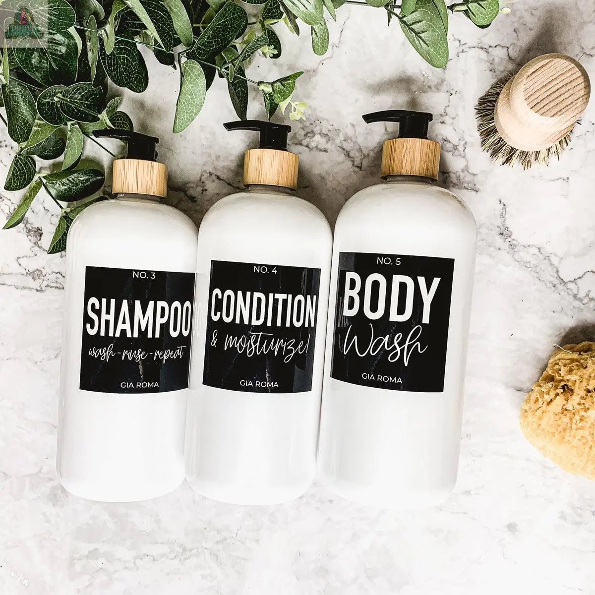 Three 32oz Plastic White Dispensers with black labels—“Shampoo,” “Conditioner & Moisturizer,” and “Body Wash”—sit on a marble surface with a plant, wooden brush, and sponge, showcasing style and sustainability.