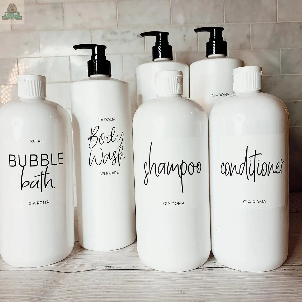 Six 32oz Plastic White Dispensers with black script labels—Bubble Bath, Body Wash, Shampoo, and Conditioner—sit on a light wood surface before tiles, offering a stylish, eco-friendly, and refillable solution.