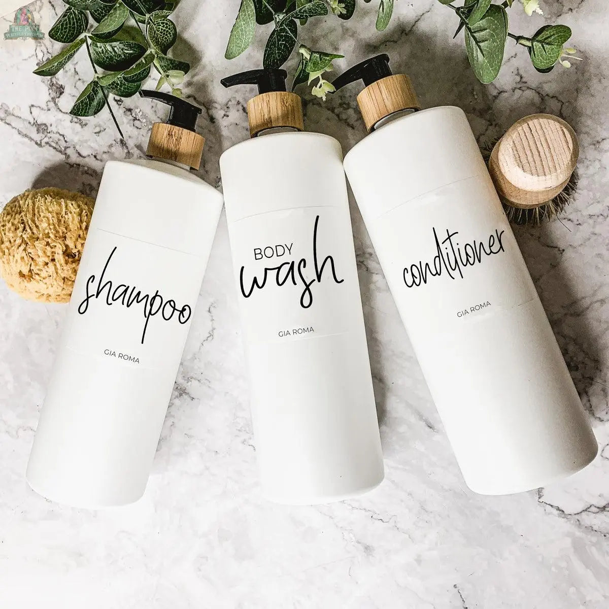 Three 32oz Plastic White Dispensers with wood pump tops and waterproof labels for shampoo, body wash, and conditioner sit on marble, surrounded by greenery and a natural sponge.