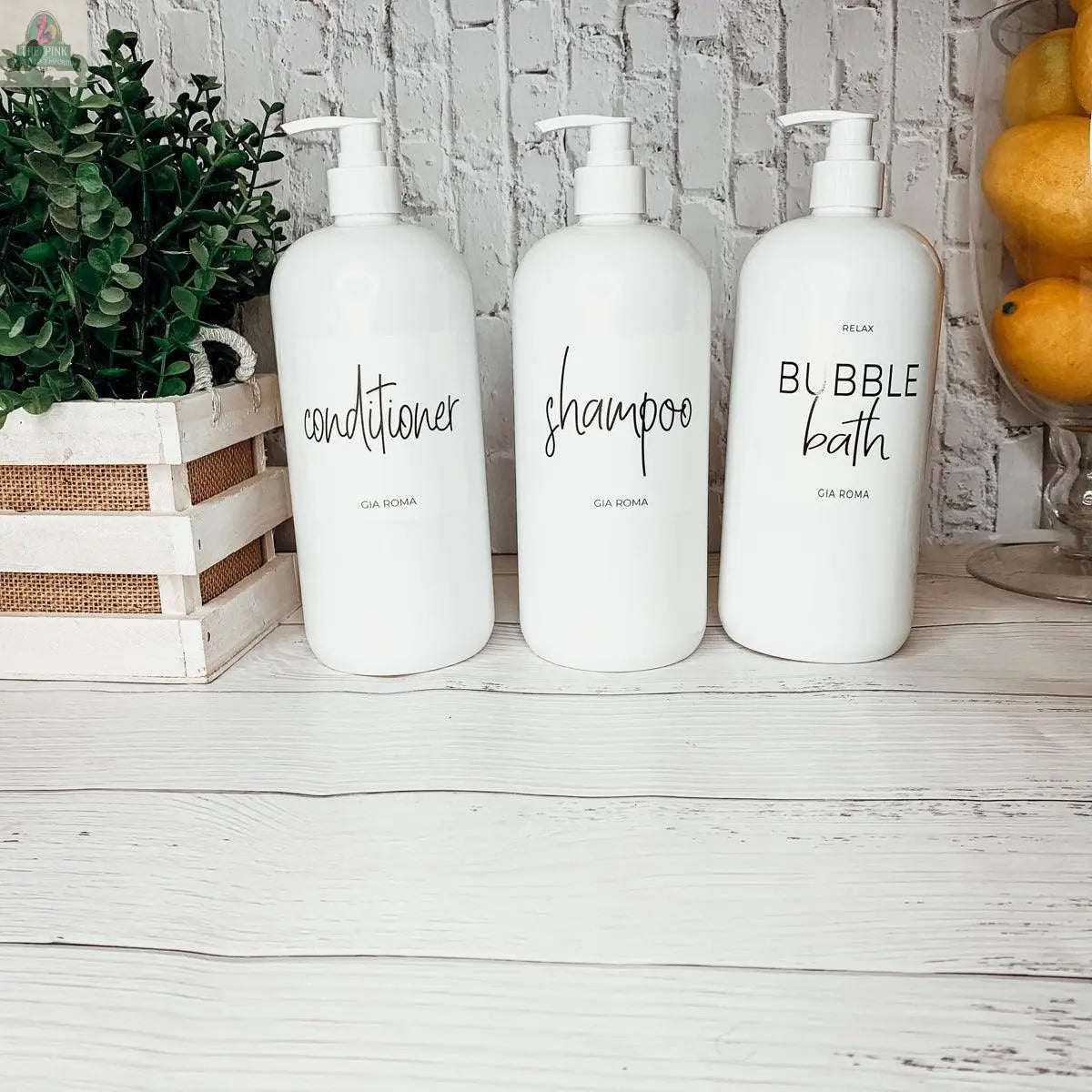 Three 32oz Plastic White Dispensers labeled conditioner, shampoo, and bubble bath sit on a white wood surface. Nearby, a green potted plant and a basket of lemons add charm against a white brick wall background.
