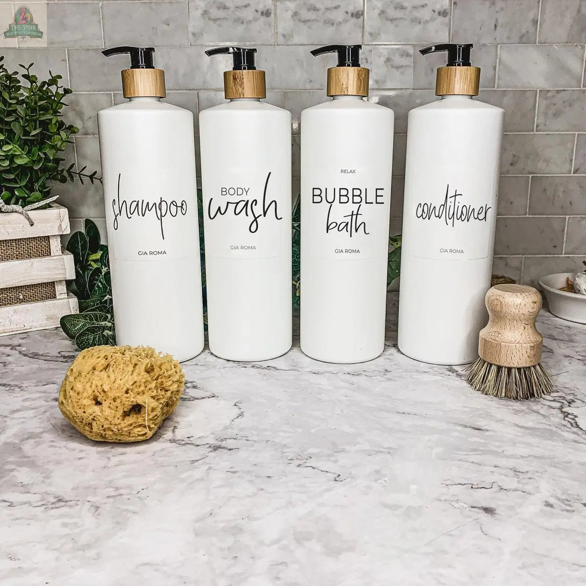 Four 32oz Plastic White Dispensers labeled shampoo, body wash, bubble bath, and conditioner sit on a marble counter with a sponge, scrub brush, and green plants—ideal for adding stylish organization to your home decor.