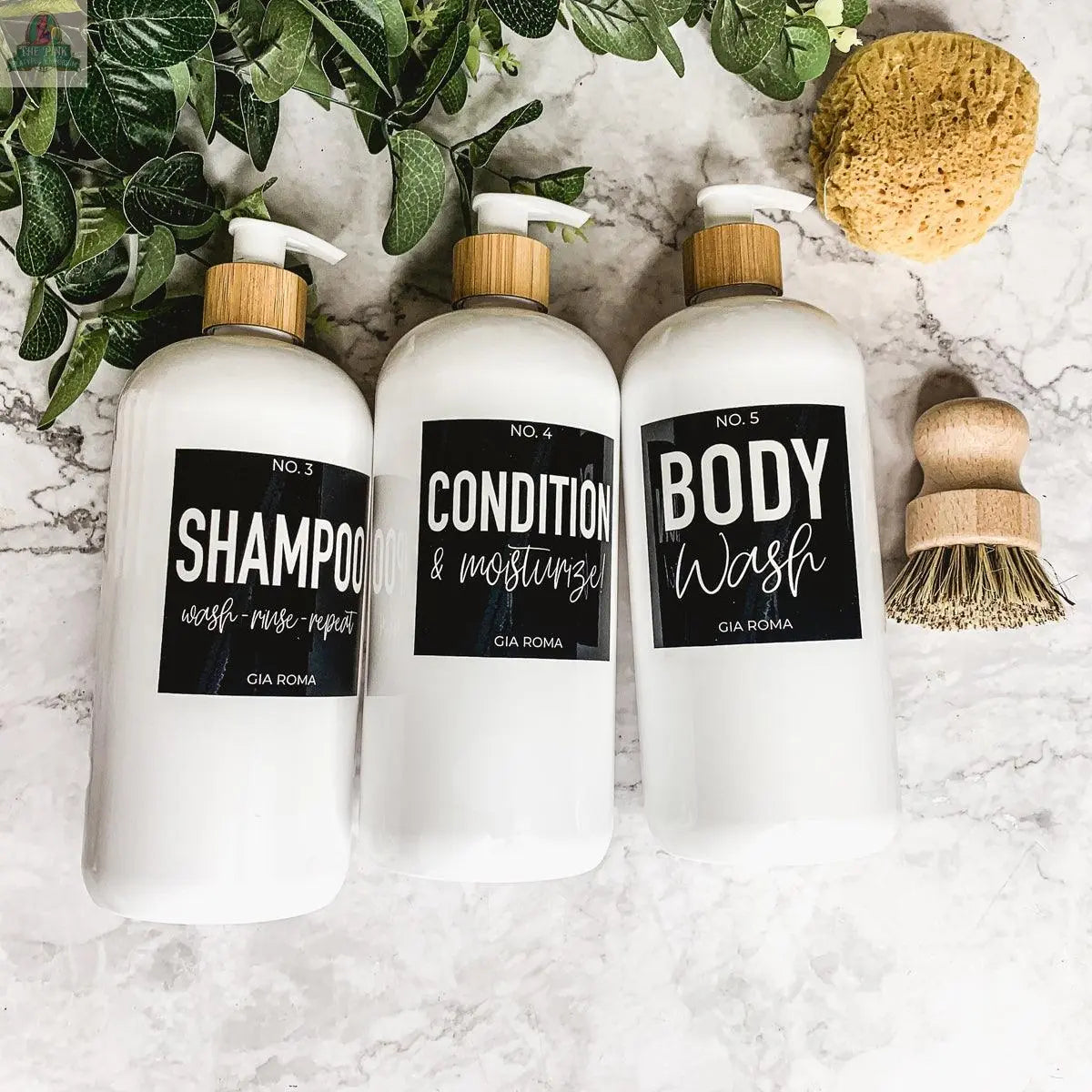 Three 32oz Plastic White Dispensers labeled “Shampoo,” “Condition,” and “Body Wash” sit on a marble surface, surrounded by green leaves, a natural sponge, and a scrub brush for a fresh, spa-like look.