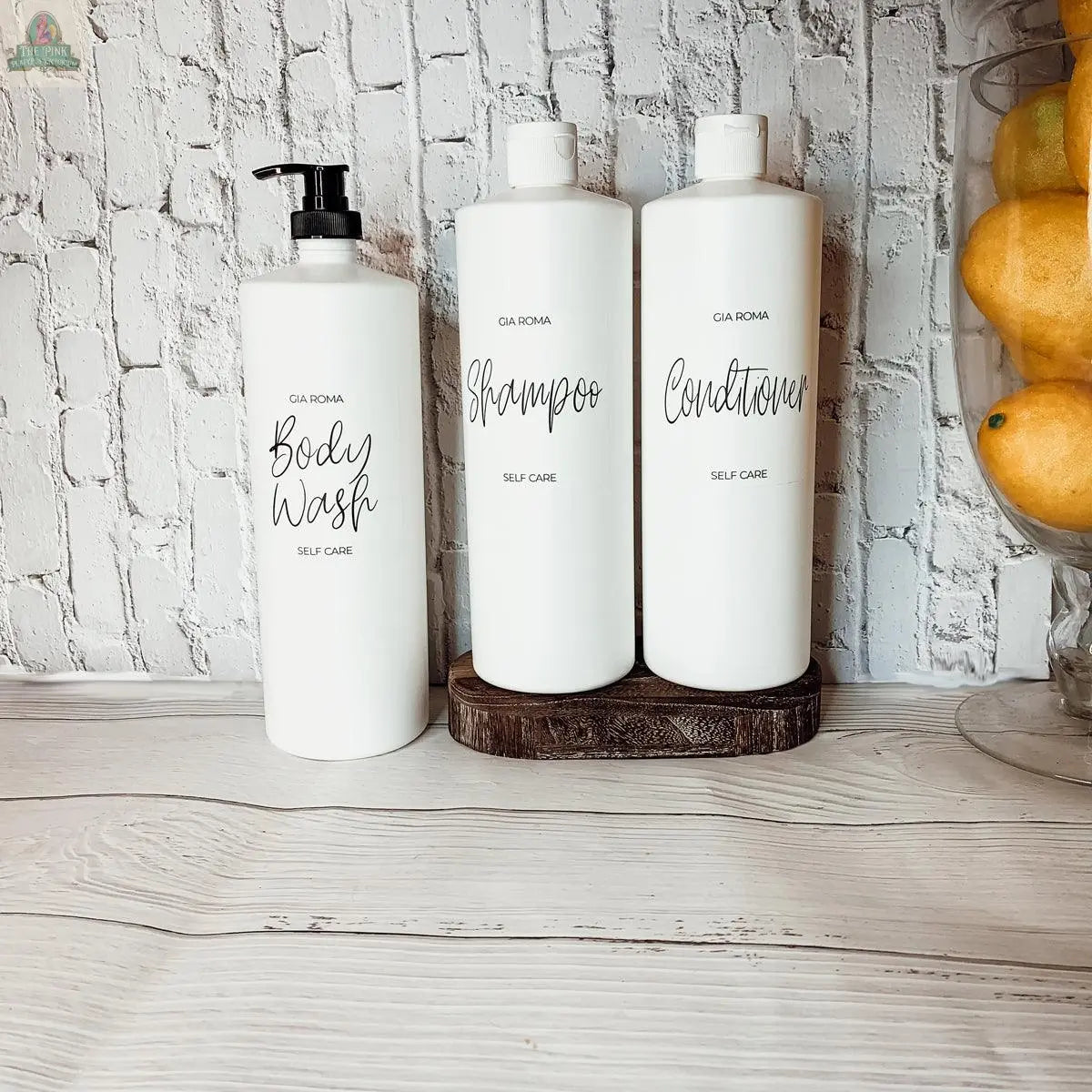 Three 32oz Plastic White Dispensers labeled Body Wash, Shampoo, and Conditioner are arranged on a light wooden surface, set against a white brick wall. A bowl of lemons to the right adds a fresh accent to the scene.