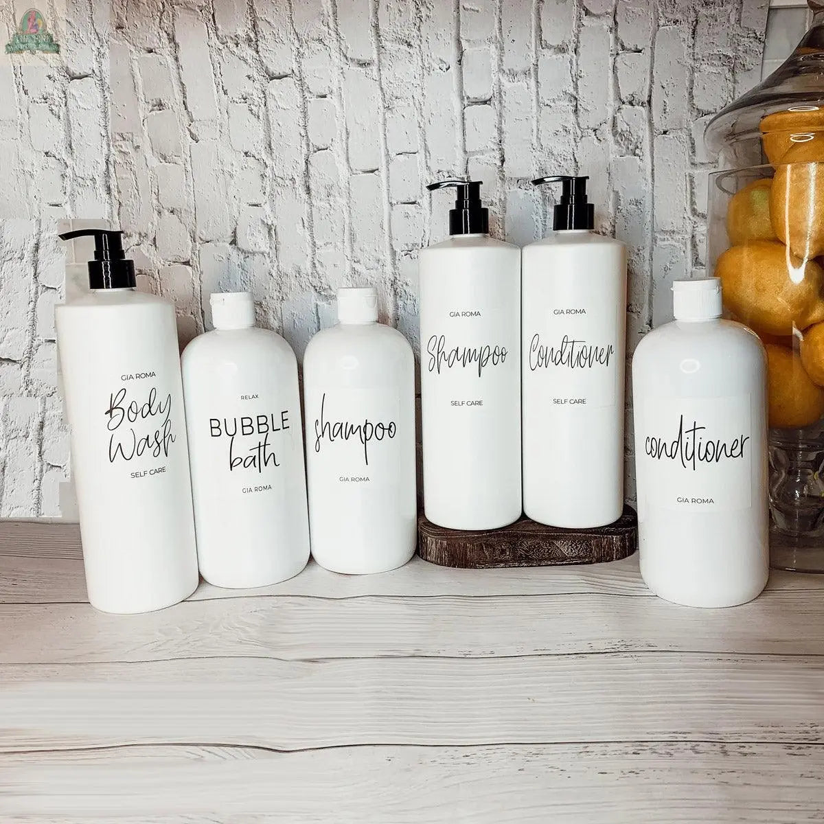 Six 32oz Plastic White Dispensers with black pumps, labeled for body wash, bubble bath, shampoo, and conditioner, are stylishly arranged as chic home decor on a white wooden surface against a white brick wall.