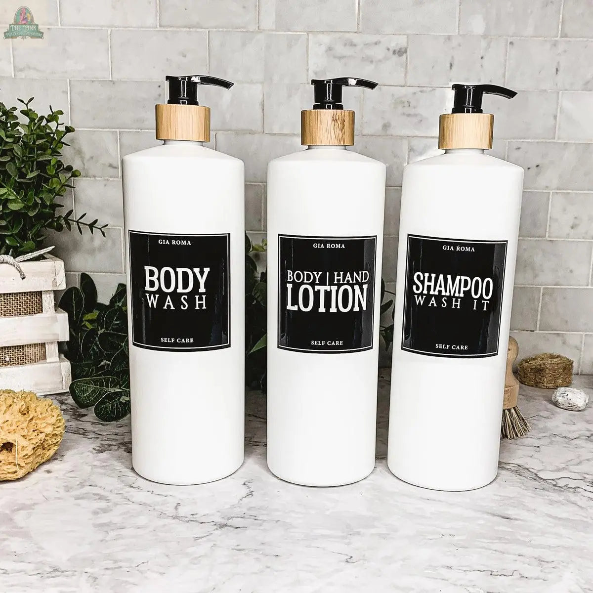 Three 32oz Plastic White Dispensers labeled Body Wash, Body | Hand Lotion, and Shampoo sit on a marble countertop with plants and bathroom accessories, adding a stylish touch to your home décor.