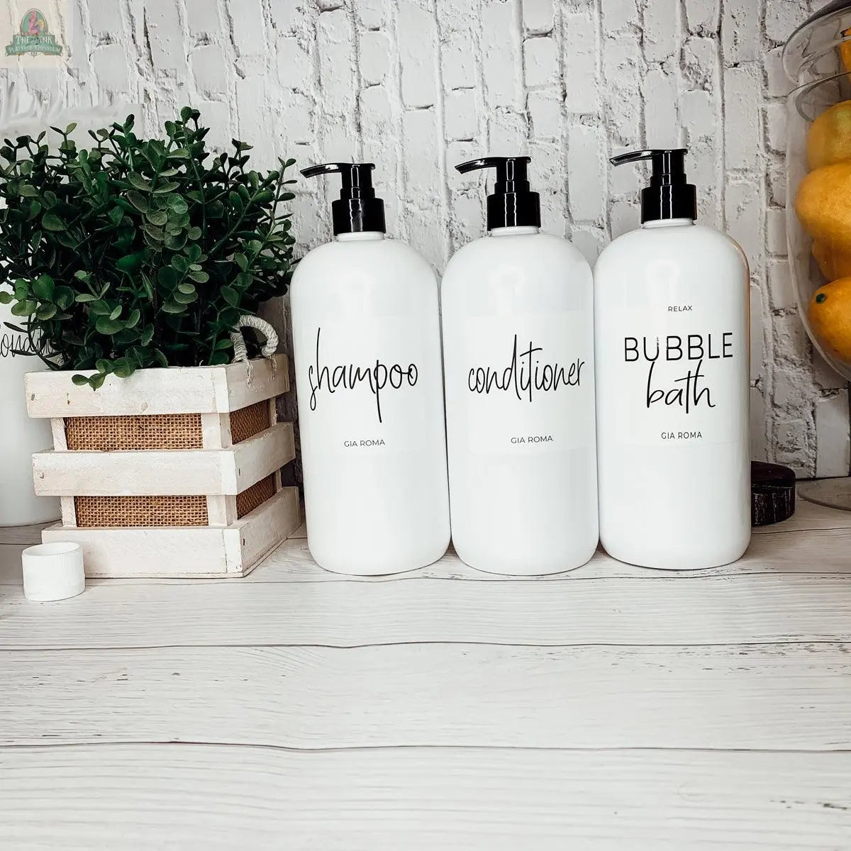 Three 32oz Plastic White Dispensers labeled shampoo, conditioner, and bubble bath rest on a white countertop next to a potted green plant in a wooden crate. Lemons in the background add a fresh accent to your home decor.