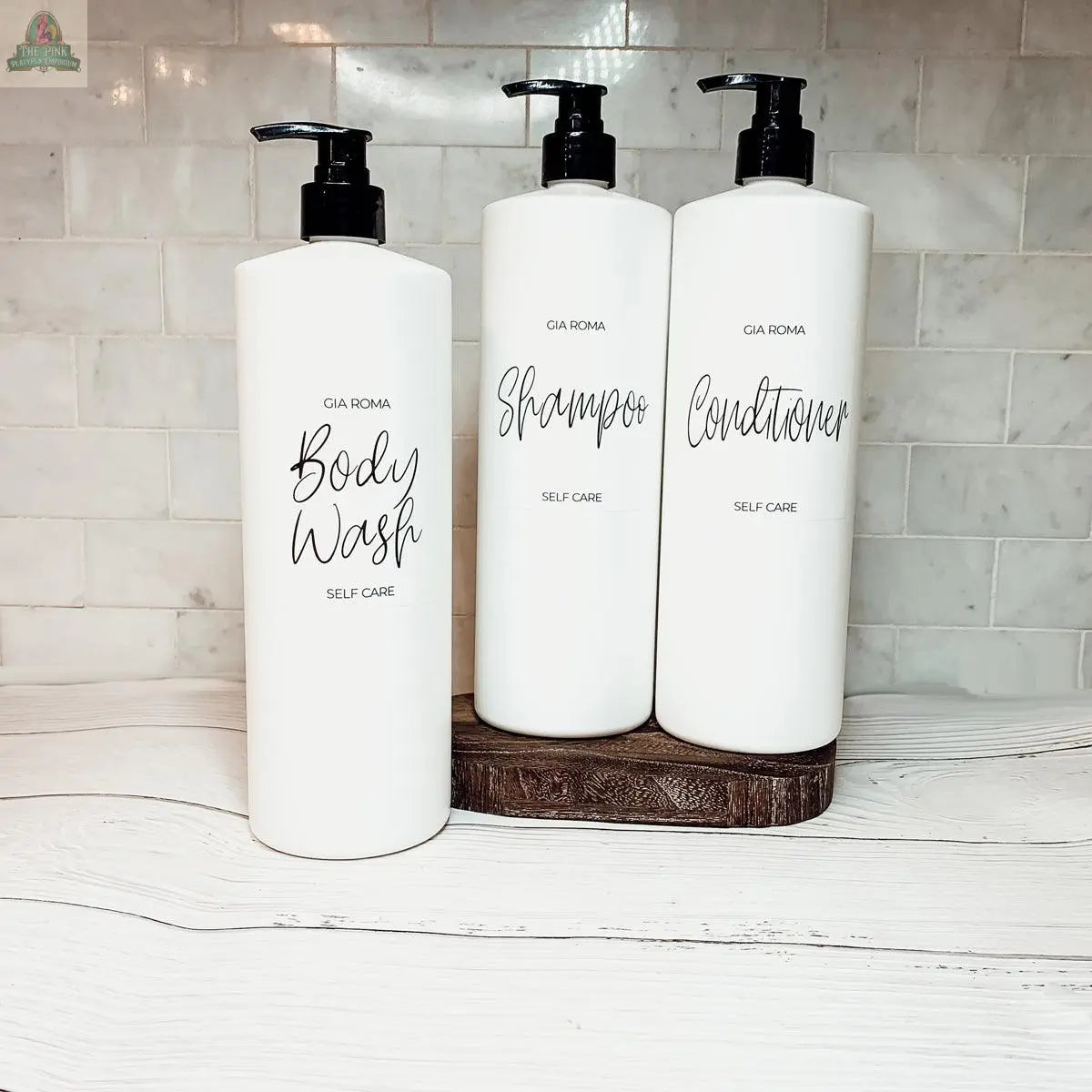Three 32oz Plastic White Dispensers labeled Body Wash, Shampoo, and Conditioner sit on a wooden tray against a white tile backsplash, creating a sleek accent for your home decor on a light-colored countertop.
