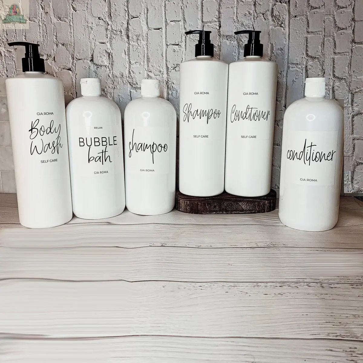 Six 32oz Plastic White Dispensers with black pump tops—labeled body wash, bubble bath, shampoo, and conditioner—are arranged on a light wood surface against a white brick wall, elevating your home decor with modern style.