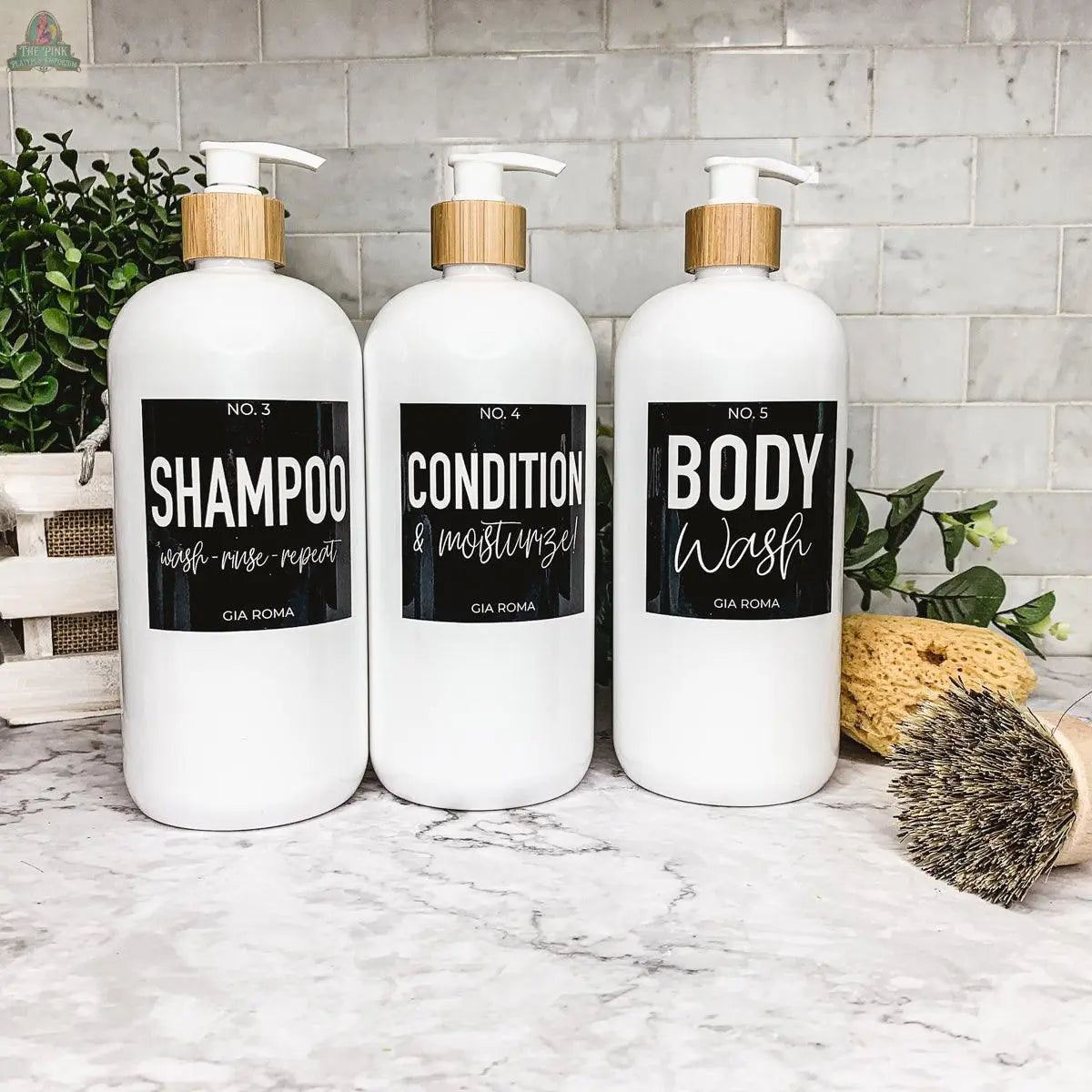 Three 32oz Plastic White Dispensers labeled Shampoo, Conditioner, and Body Wash sit on a marble counter with a plant, brush, sponge, and basket—ideal for adding stylish, sustainable home decor.