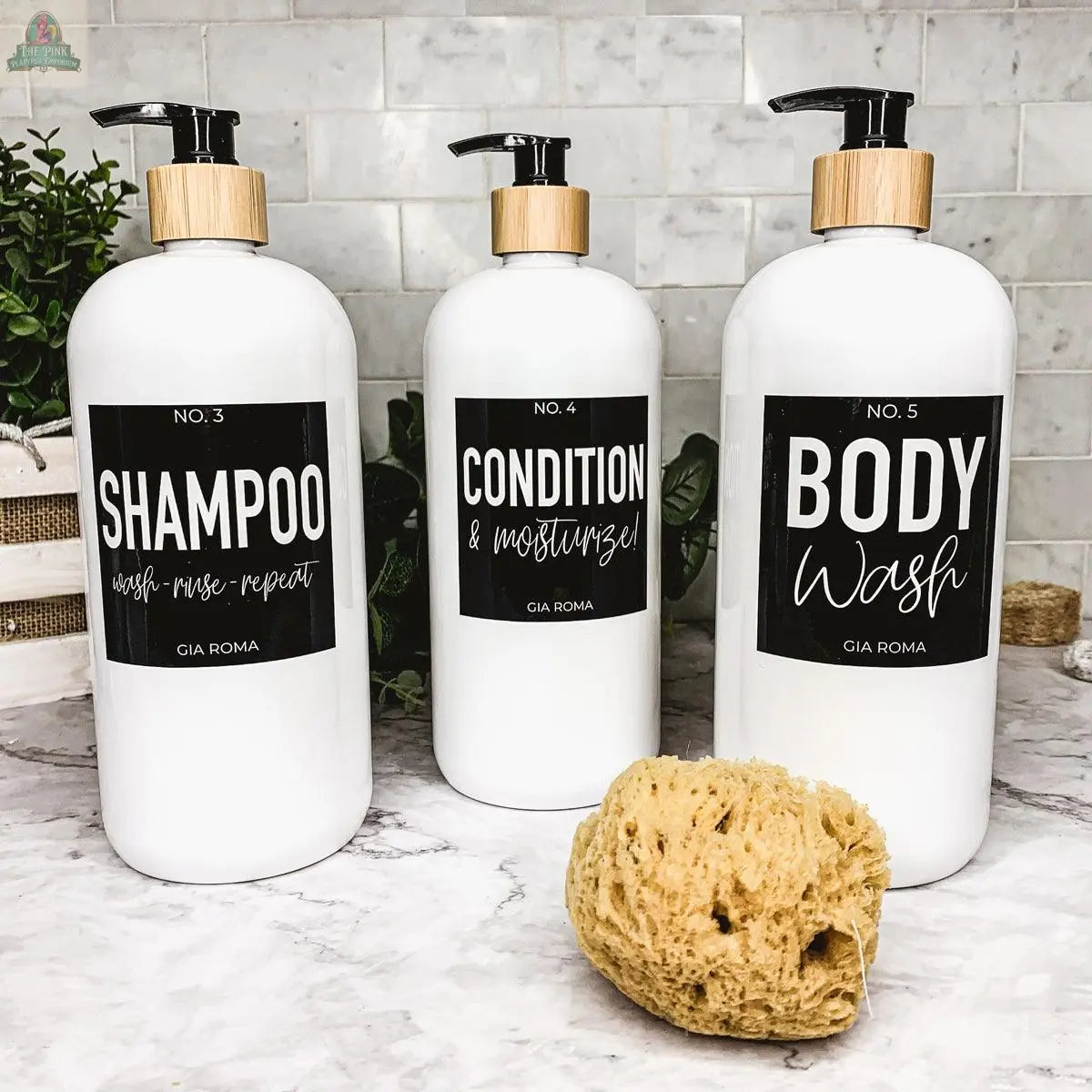Three 32oz Plastic White Dispensers labeled Shampoo, Conditioner & Moisturize!, and Body Wash sit on a marble counter with a sponge. Each features a black label and wooden pump top, adding a stylish touch to your decor. A plant completes the scene.