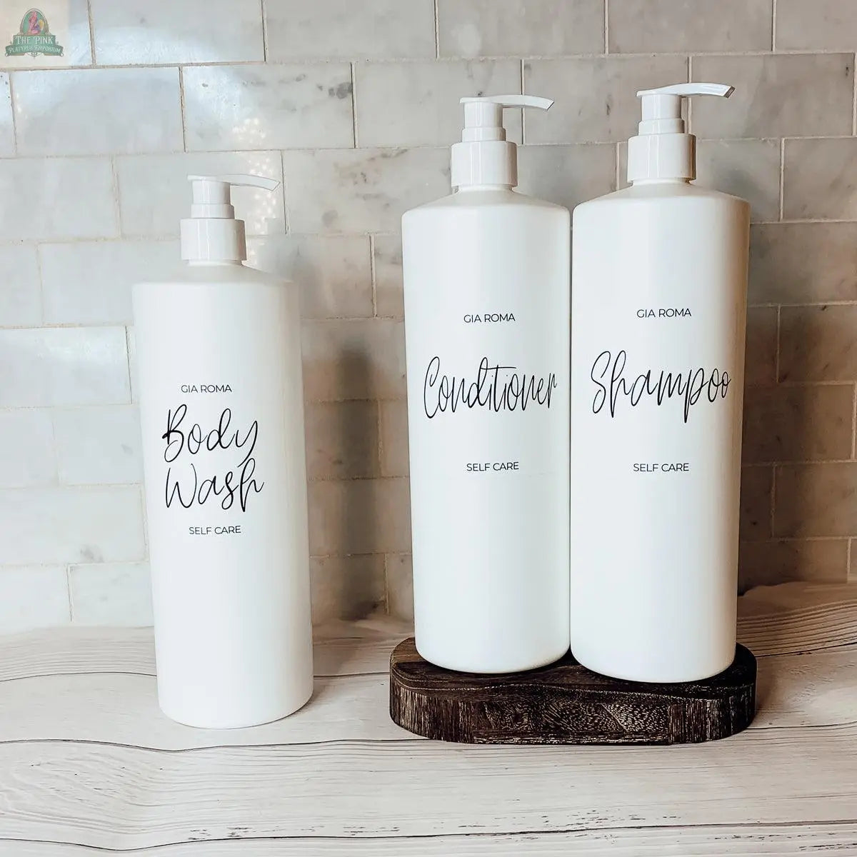 Three 32oz Plastic White Dispensers labeled Body Wash, Conditioner, and Shampoo sit on a light countertop, with the latter two on a wooden stand against a tiled wall, bringing a touch of elegance to your home decor.