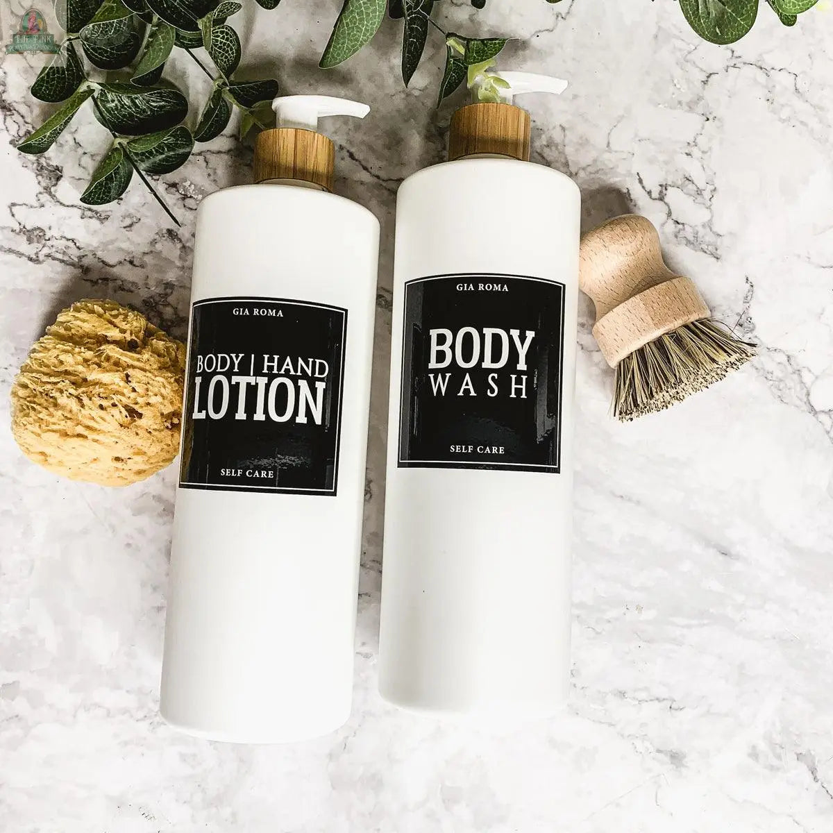Two 32oz Plastic White Dispensers labeled Body Hand Lotion and Body Wash with waterproof labels feature wooden pumps, shown on marble next to a yellow sponge, wooden scrub brush, and green plant leaves.