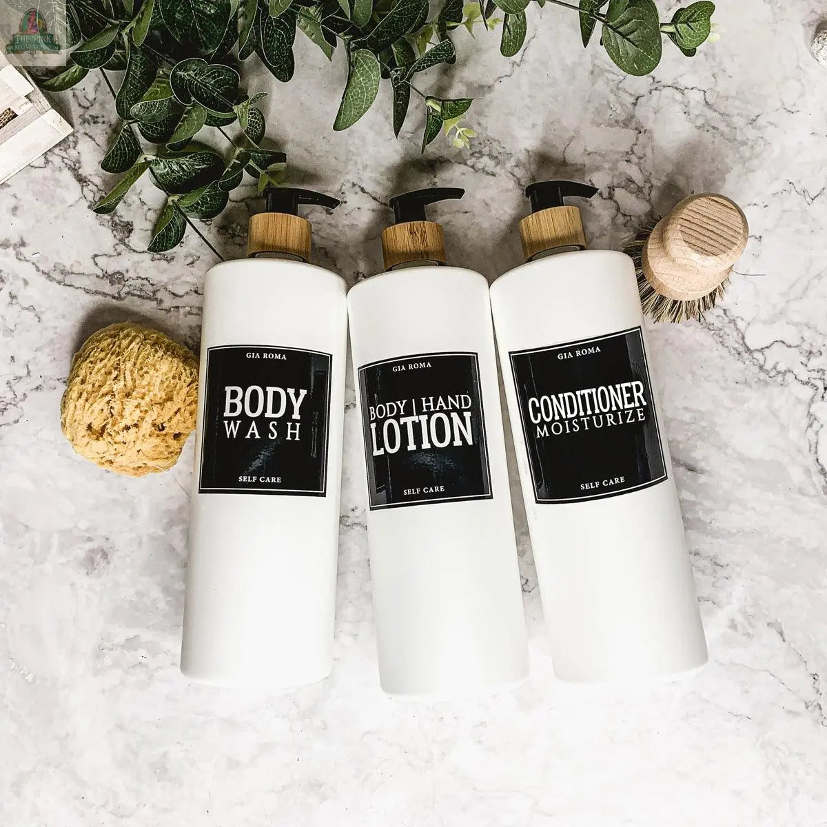 Three 32oz Plastic White Dispensers labeled Body Wash, Hand Lotion, and Conditioner Moisturize sit on a marble surface with green leaves, a sponge, and a wooden brush—perfect for stylish home decor.