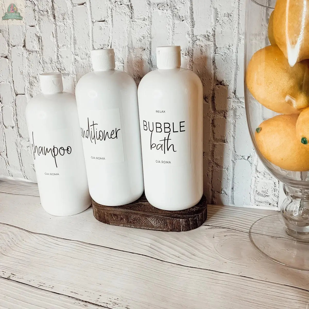 Three 32oz Plastic White Dispensers labeled shampoo, conditioner, and bubble bath sit on a wooden tray by a glass bowl of lemons against a white brick wall—ideal for stylish, eco-friendly home decor.