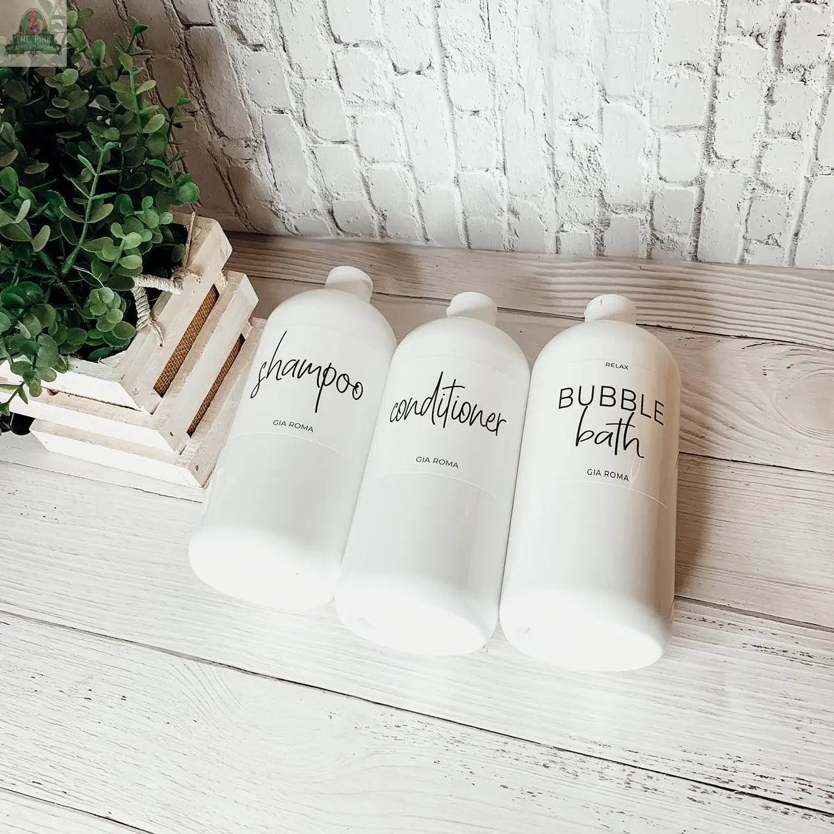 Three 32oz Plastic White Dispensers labeled “shampoo,” “conditioner,” and “bubble bath” sit on a white wood surface next to a green potted plant, bringing homey style to a white brick wall backdrop.