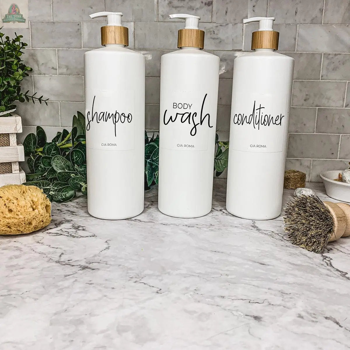 Three 32oz Plastic White Dispensers labeled "shampoo," "body wash," and "conditioner" sit on a marble countertop with green plants, a sponge, and a brush nearby, bringing style and sustainability to your home decor.