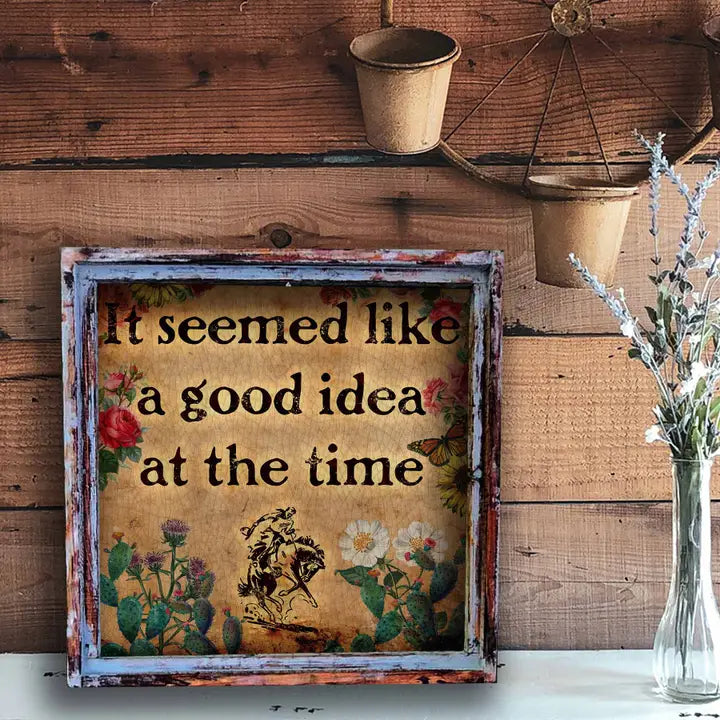 Decorative sign with "it seemed like a good idea at the time" text and floral design on a wooden wall.