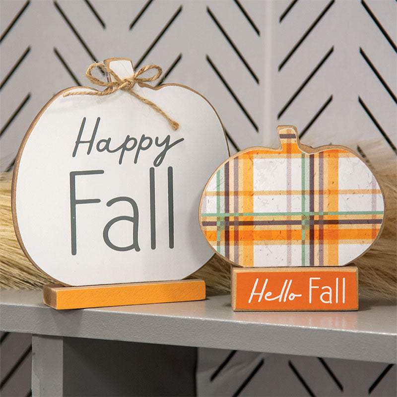 Two decorative autumn pumpkin signs on a shelf. The larger left sign is white with 'Happy Fall' in dark gray script and block lettering, with a small jute bow and orange base.