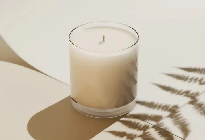 The MAHALO - *Signature Series* candle by Moon Water Apothecary, in a clear glass jar, sits on a light surface with a fern leaf shadow nearby, filling the minimalist space with its sweet, earthy Hawaiian Koa Wood exclusive blend.