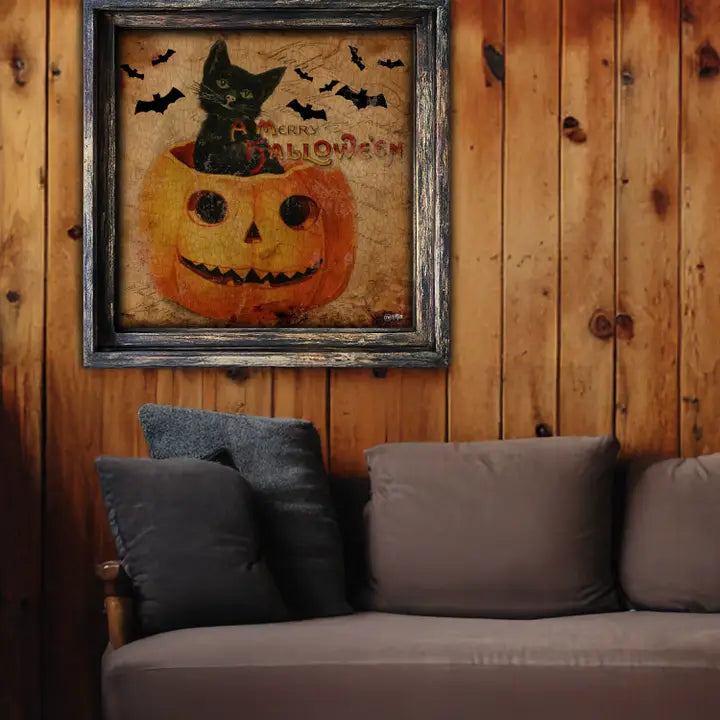Halloween-themed framed picture of a black cat in a pumpkin on a wooden wall above a gray sofa.