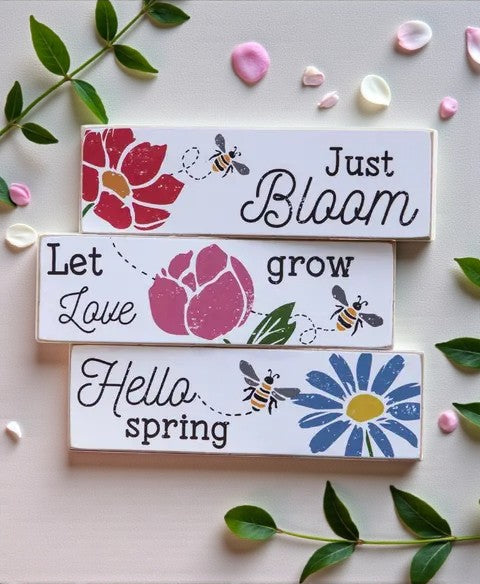 Decorative wooden signs with floral designs and text on a light background with flowers and leaves.