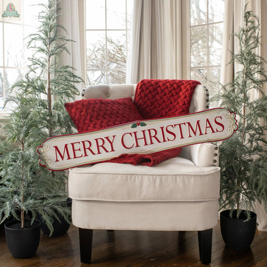 A white armchair with red knit pillows and a blanket sits between two potted pine trees. The 40" RED & WHITE MERRY CHRISTMAS PLAQUE is placed across the chair, while large windows in the background enhance the holiday décor.