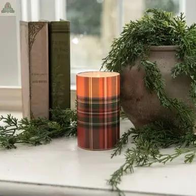 A 5" RED TARTAN 3D FLAME GLASS CANDLE with a plaid design is displayed on a white surface beside a ceramic planter with greenery and two vintage books, surrounded by additional greenery near a window.